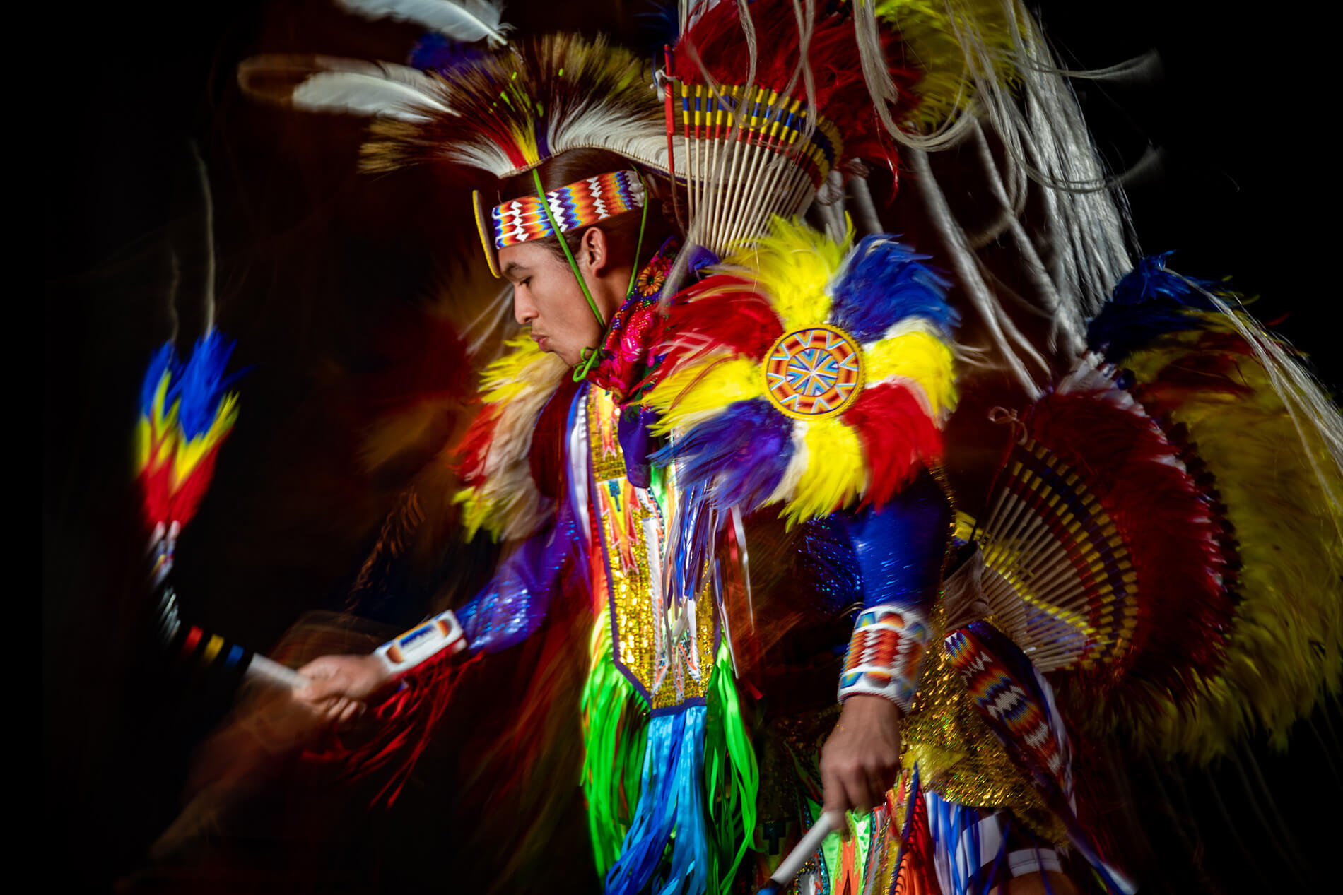 A Native American man  wearing a bright blue, yellow, and red regalia dancing in front of a black backdrop.