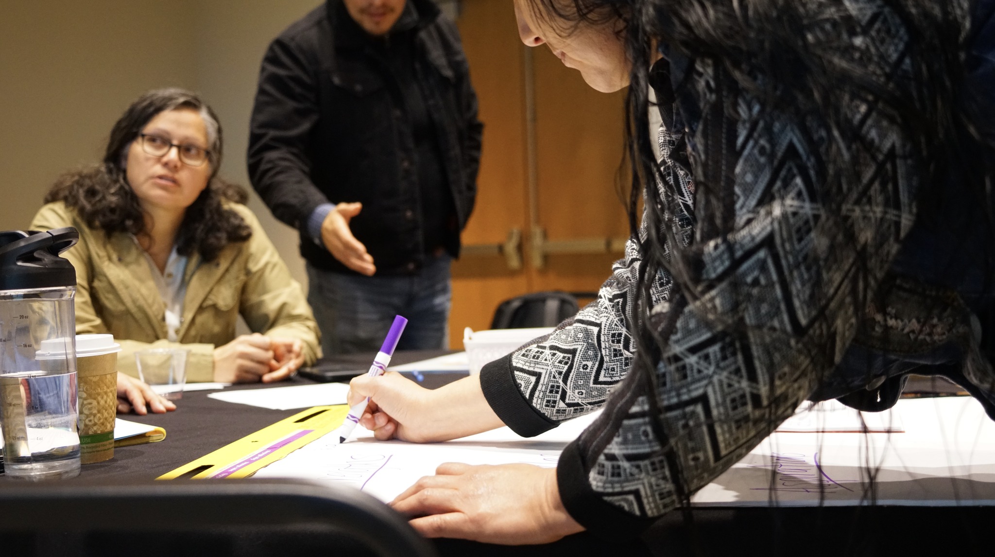 A Native American woman in a  black and white sweater writing notes on a poster board with a purple marker.