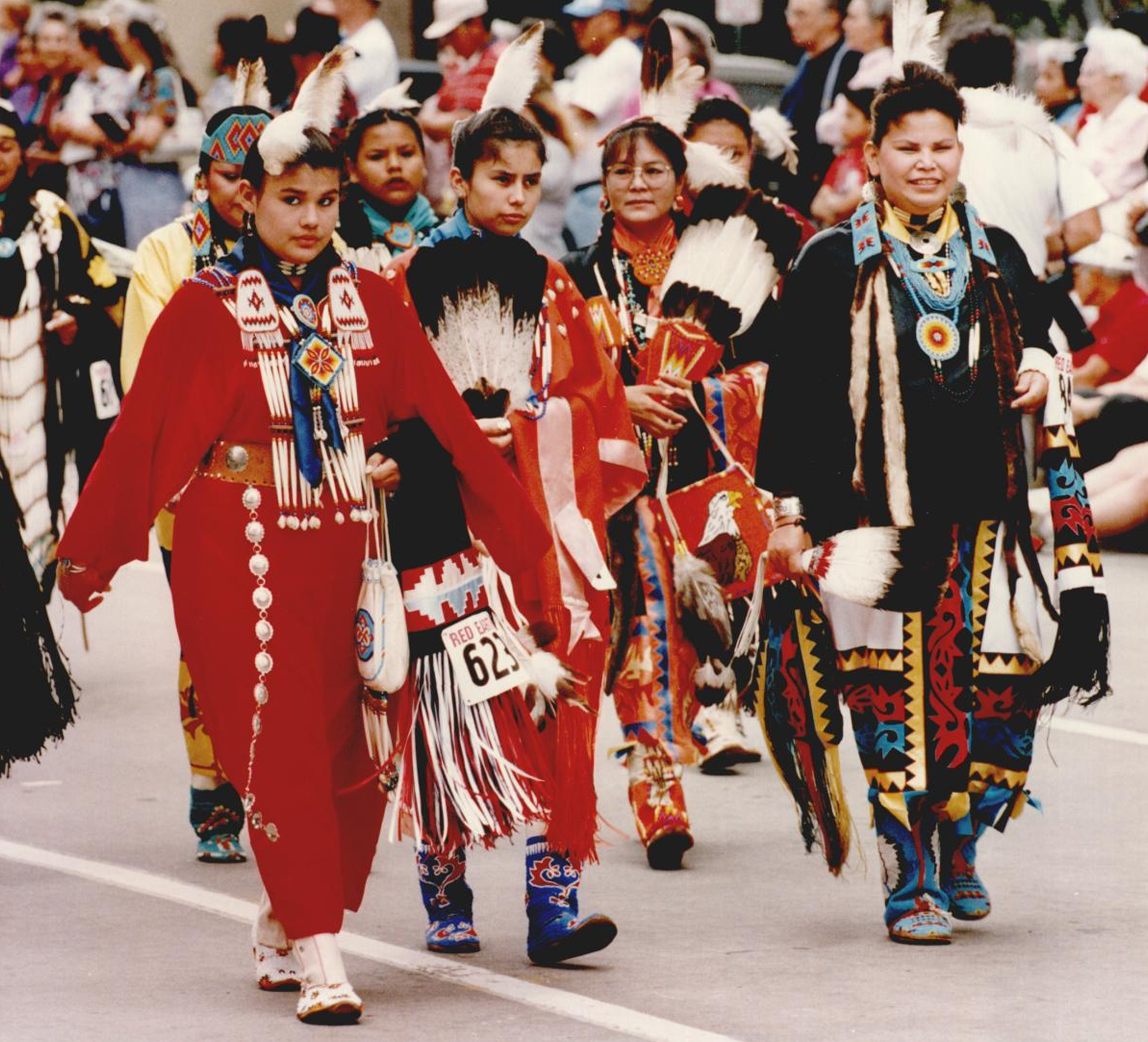 Youth wearing tribal dance regalia walking down the street as part of a parade.