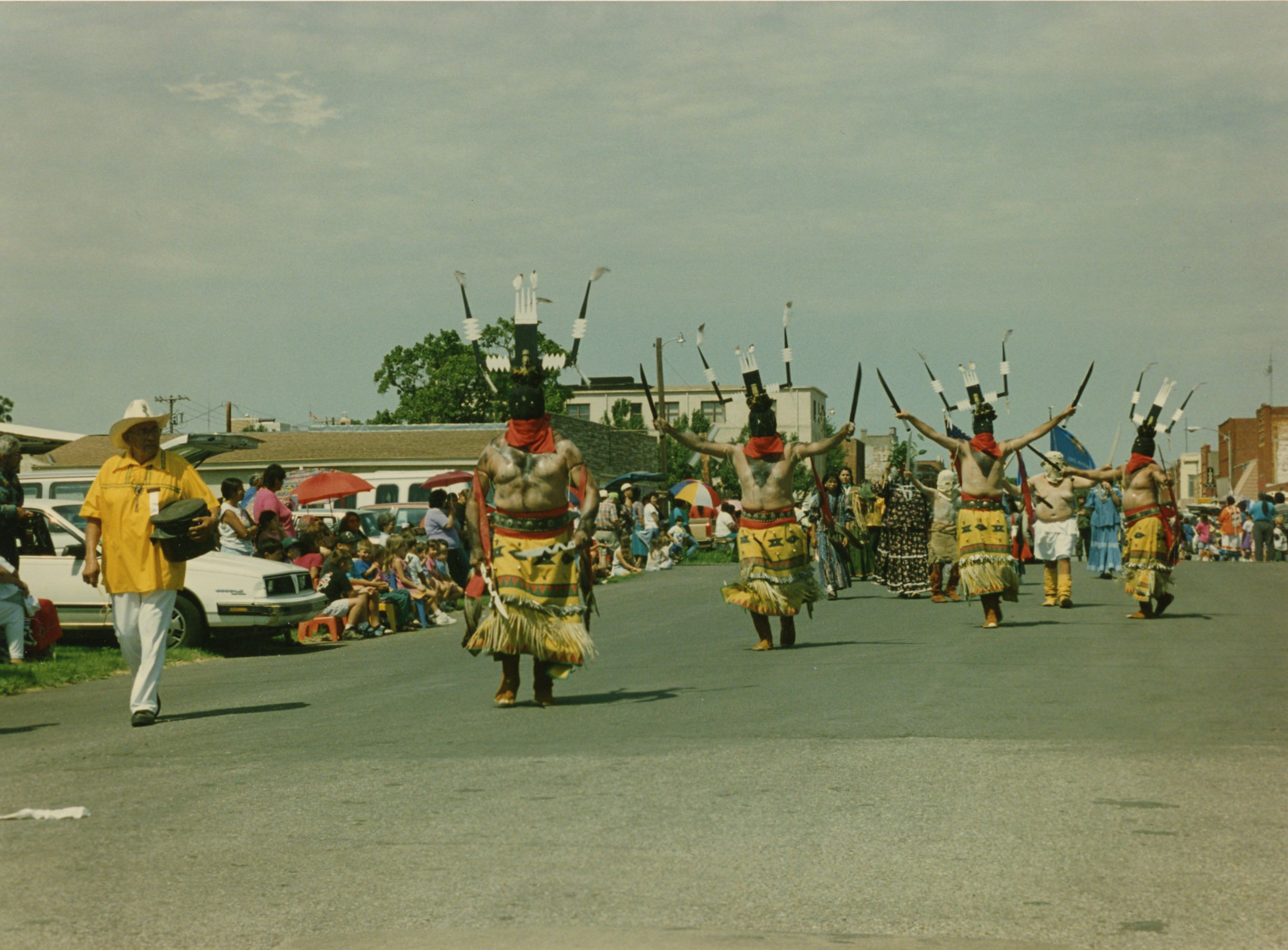 Men in tribal regalia walking down a street as part of a procession.