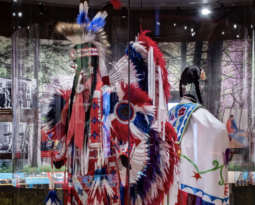 Male and Female red, white, and blue regalia behind glass inside the "Regrowth" section of the OKLA HOMMA Exhibit at First Americans Museum.