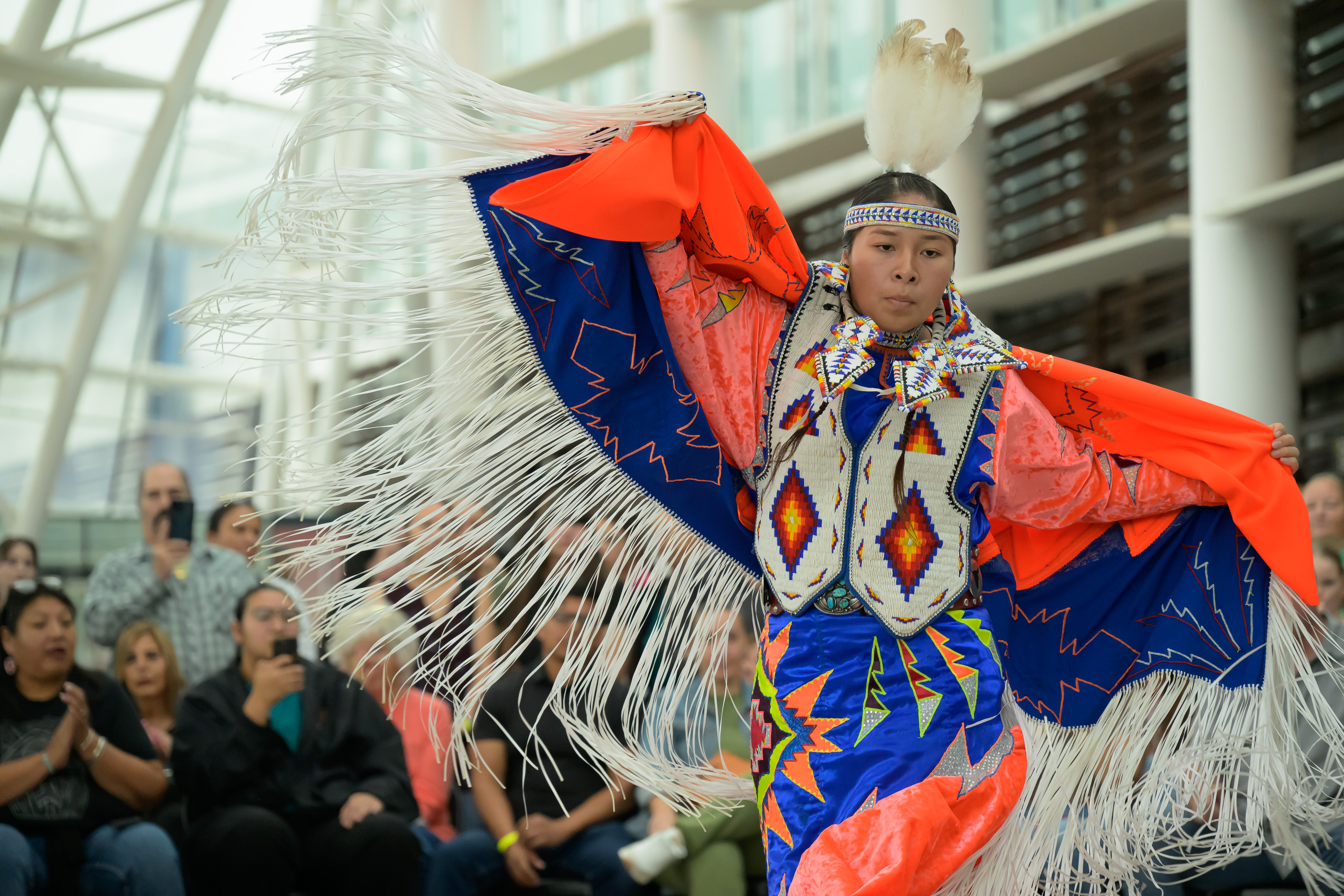 A Native American dancer wearing bright blue, orange, and white regalia with beaded patterns and a shawl with long white fringe performs before an audience in the Hall of The People during First Americans Museum’s 2025 Indigenous Peoples’ Day celebration.