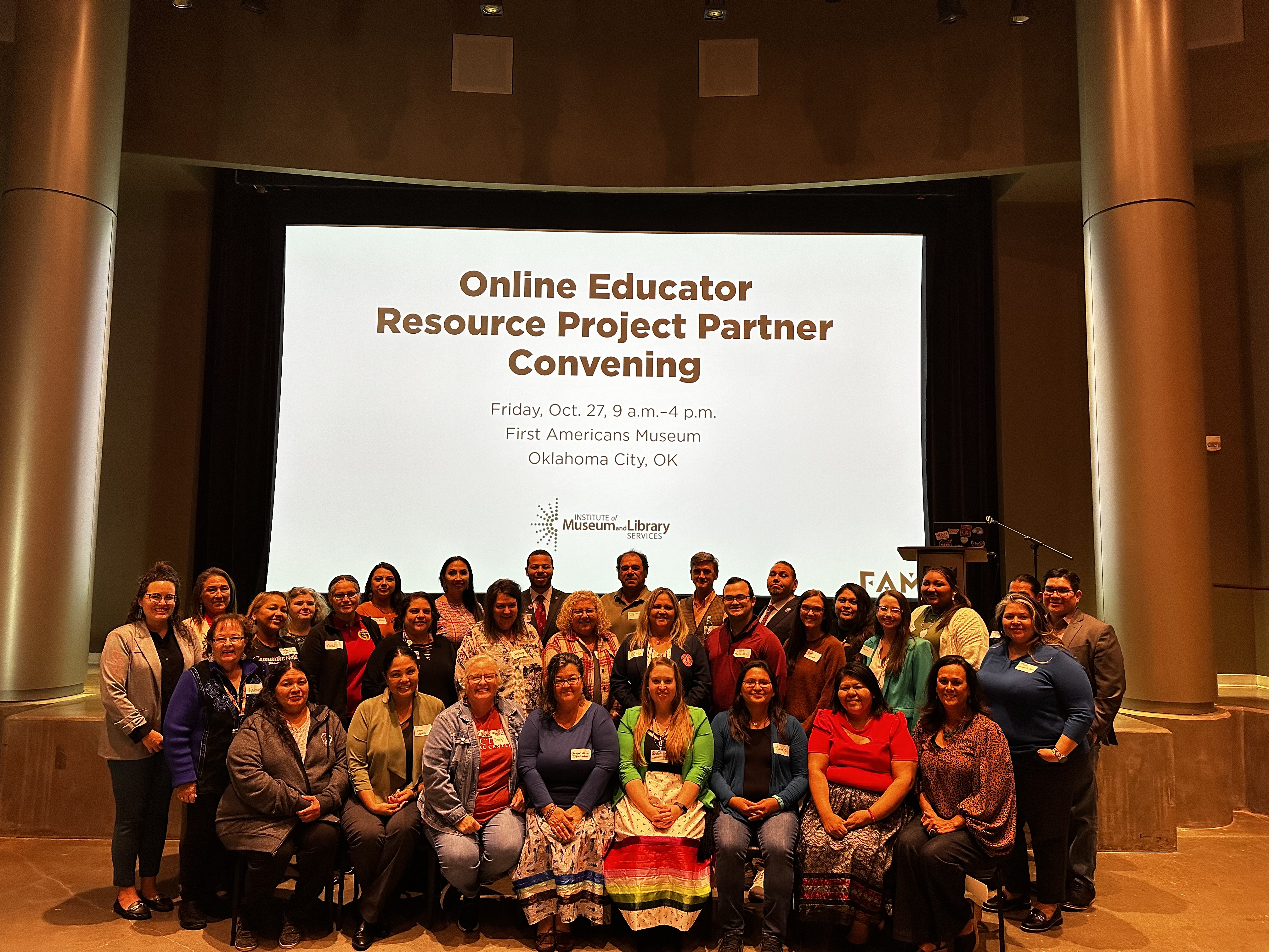 A group of people facing the camera, behind them is a projector screen that reads "Online Educator Resource Project Partner Convening, Friday, Oct. 27, 9 a.m.-4 p.m. First Americans Museum, Oklahoma City, OK." 