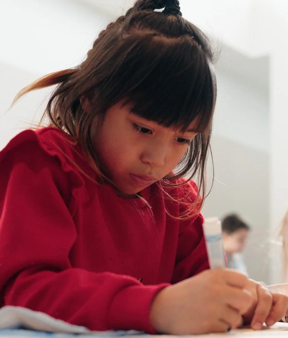 A young Native American girl in a red sweater writing with a blue marker. 