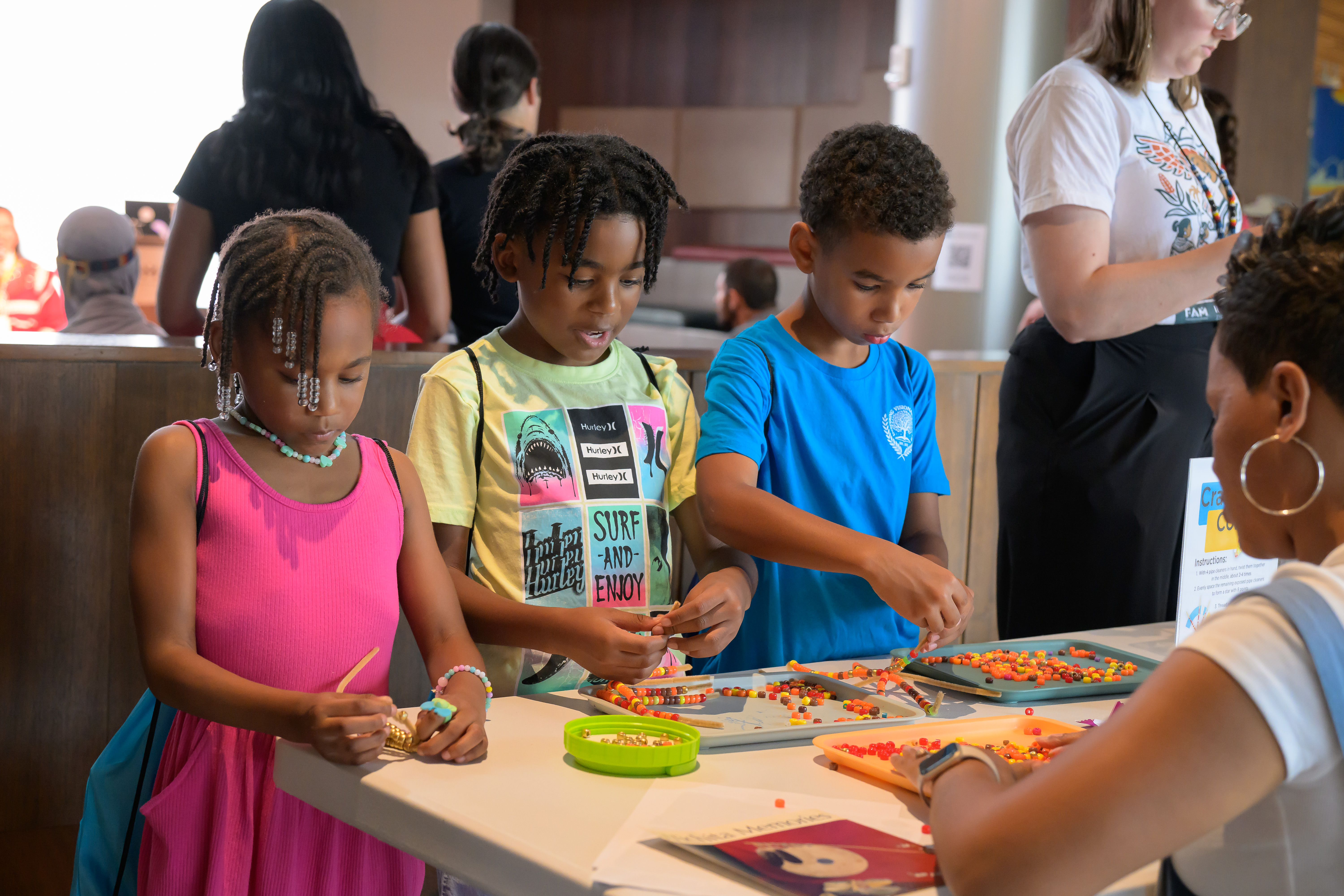 Three young children stand at a table creating ears of corn from pipe cleaners and yellow, orange, and brown beads during First Americans Museum’s 2025 Indigenous Peoples’ Day celebration.