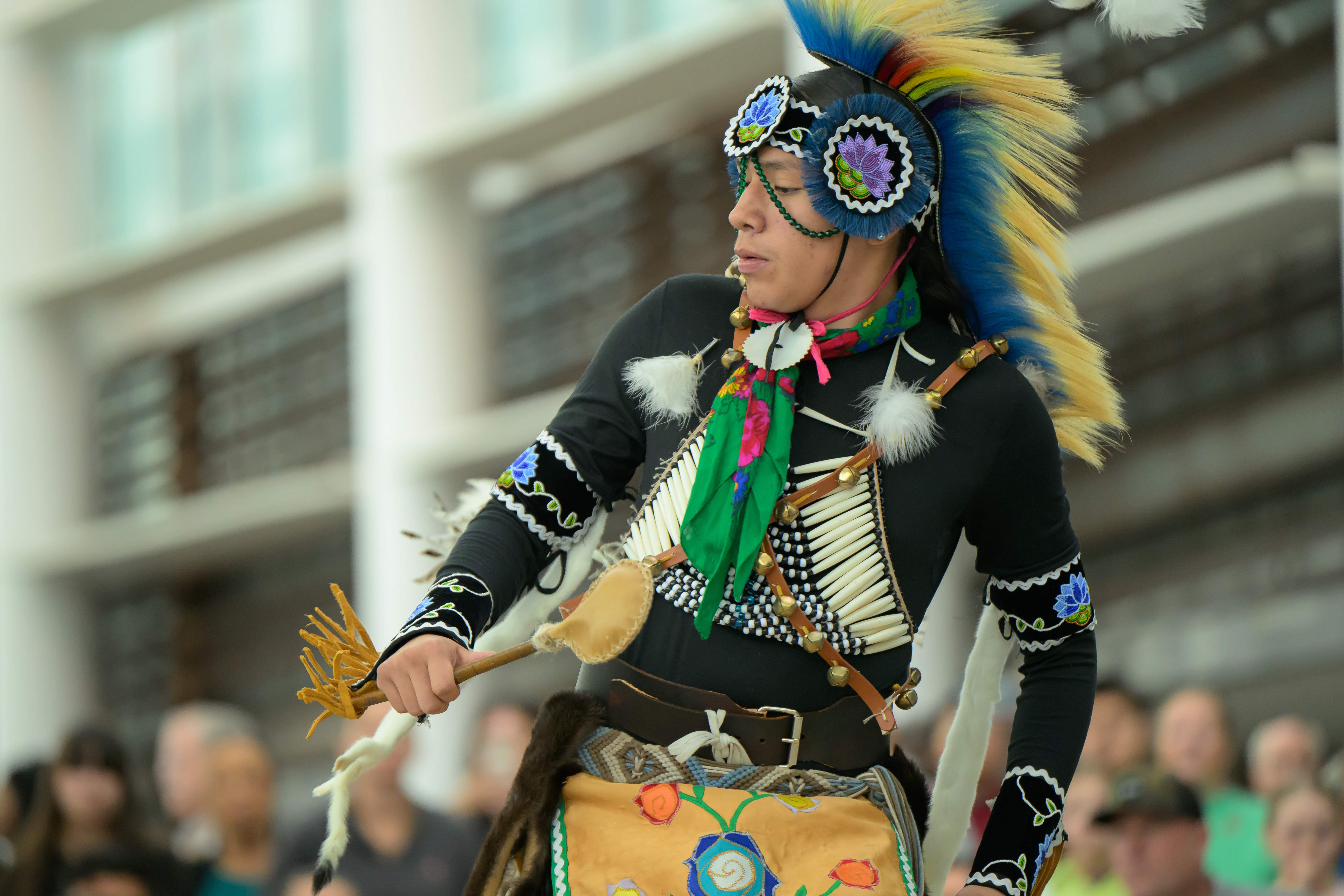A Native American man in black, white, and blue regalia  dances with a rattle in the Hall of the People at First Americans Museum's 2025 Indigenous Peoples'
 Day celebration.